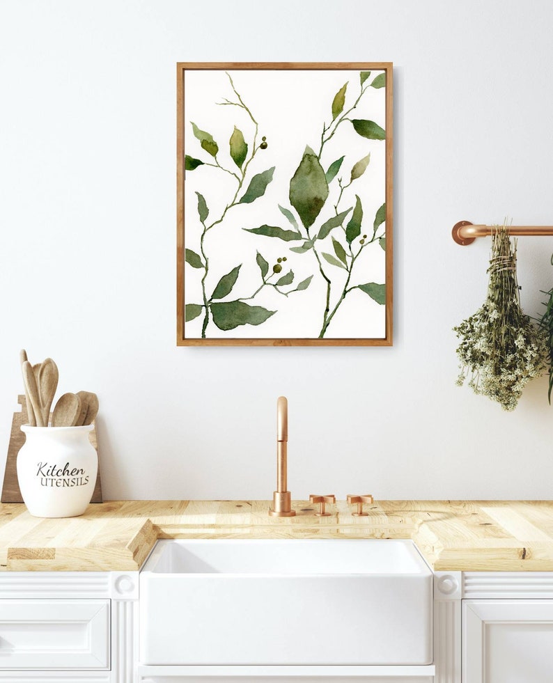 Puede incluir: Una pintura de acuarela enmarcada de hojas verdes y ramas sobre un fondo blanco. La pintura est&aacute; colgada en una pared blanca sobre un fregadero de cocina con un grifo de cobre.