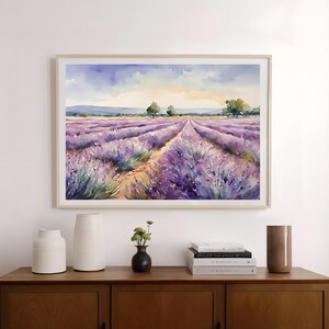 May include: A watercolor painting of a lavender field with a dirt path leading through the rows of purple flowers. The painting is framed in a light wood frame and is hanging on a white wall.