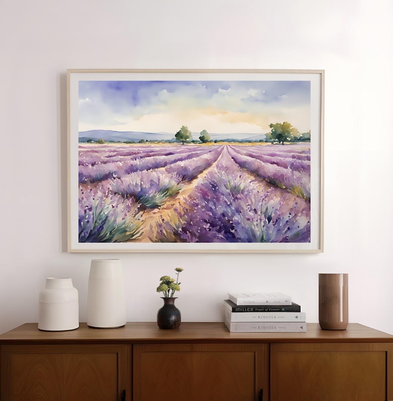 May include: A watercolor painting of a lavender field with a dirt path leading through the rows of purple flowers. The painting is framed in a light wood frame and is hanging on a white wall.
