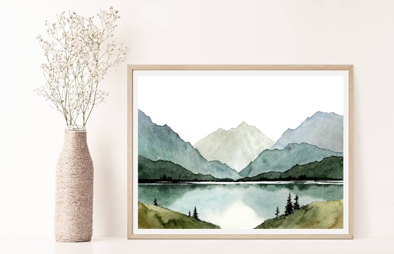 May include: Watercolor painting of a mountain range with a lake in the foreground. The mountains are painted in shades of blue and green, and the lake is a light blue. The painting is framed in a light wood frame.