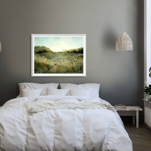 May include: A watercolor painting of a field of wildflowers with a forest in the background. The painting is framed in a white frame and hangs on a gray wall above a white bed.