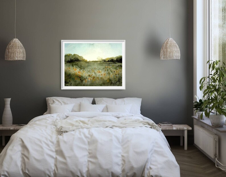 May include: A watercolor painting of a field of wildflowers with a forest in the background. The painting is framed in a white frame and hangs on a gray wall above a white bed.
