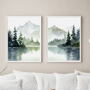 May include: Two watercolor paintings of a misty mountain lake scene with evergreen trees. The paintings are framed in white and hung on a white wall.