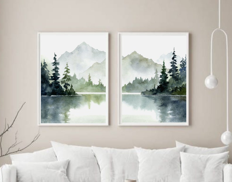 May include: Two watercolor paintings of a misty mountain lake scene with evergreen trees. The paintings are framed in white and hung on a white wall.