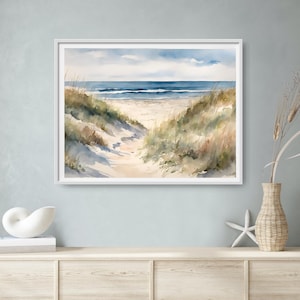 May include: A framed watercolour painting depicting a beach scene. A sandy path winds towards the sea, bordered by grassy dunes. The sky is blue with fluffy clouds. The artwork conveys a sense of peace and coastal charm.