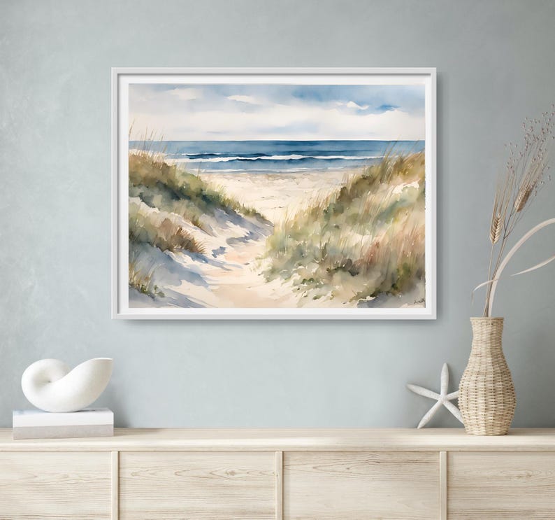May include: A framed watercolour painting depicting a beach scene. A sandy path winds towards the sea, bordered by grassy dunes. The sky is blue with fluffy clouds. The artwork conveys a sense of peace and coastal charm.
