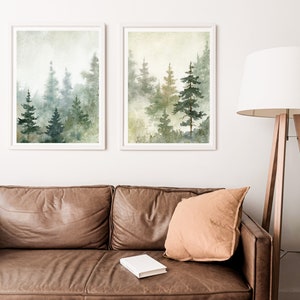 Puede incluir: Dos pinturas de acuarela enmarcadas de un bosque brumoso con altos &aacute;rboles verdes. Las pinturas est&aacute;n colgadas en una pared blanca sobre un sof&aacute; de cuero marr&oacute;n con un coj&iacute;n beige y un libro blanco.