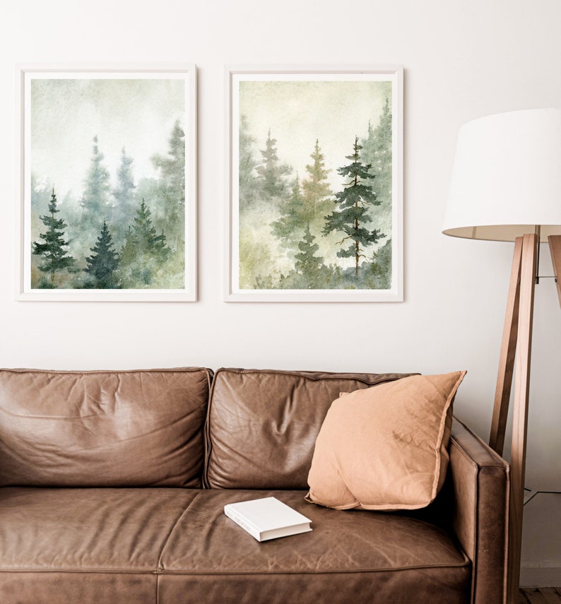 Puede incluir: Dos pinturas de acuarela enmarcadas de un bosque brumoso con altos &aacute;rboles verdes. Las pinturas est&aacute;n colgadas en una pared blanca sobre un sof&aacute; de cuero marr&oacute;n con un coj&iacute;n beige y un libro blanco.