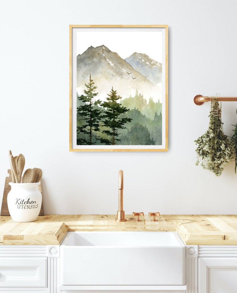 May include: A watercolor painting of a mountain landscape with two pine trees in the foreground. The mountains are a muted green and gray, and the sky is a light blue. The painting is framed in a light wood frame.