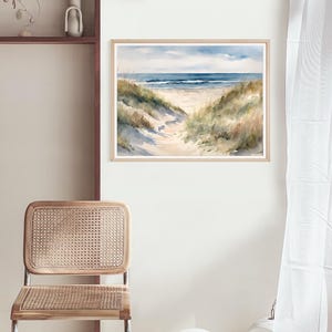 May include: Watercolour painting depicting a beach scene with a path to the sea. The artwork shows a sandy path, grassy dunes, and a blue sky with clouds. The painting is framed in a light wood frame.