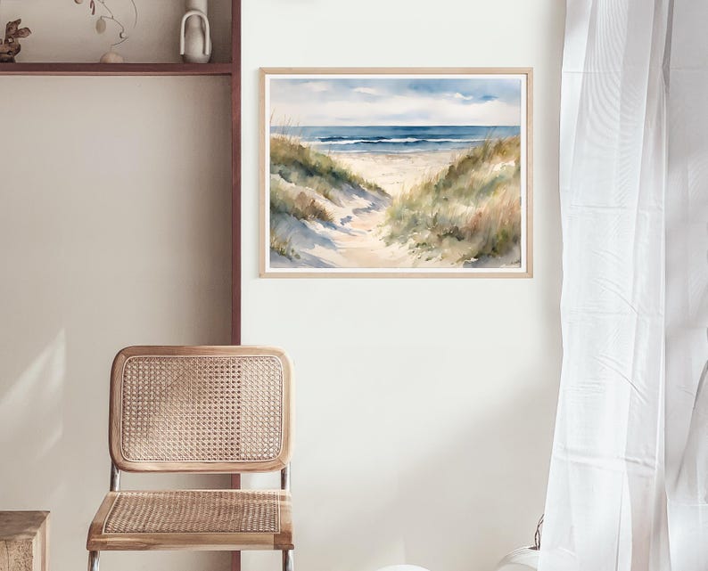 May include: Watercolour painting depicting a beach scene with a path to the sea. The artwork shows a sandy path, grassy dunes, and a blue sky with clouds. The painting is framed in a light wood frame.