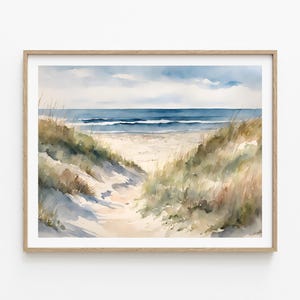 May include: Watercolour painting of a beach scene. The artwork shows a sandy path leading to the ocean, framed by grassy dunes. The sky is a soft blue with clouds, and the sea is a darker blue. The painting is in a wooden frame.