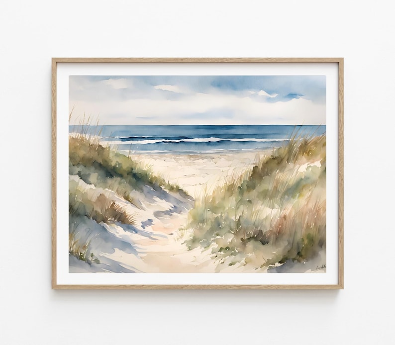 May include: Watercolour painting of a beach scene. The artwork shows a sandy path leading to the ocean, framed by grassy dunes. The sky is a soft blue with clouds, and the sea is a darker blue. The painting is in a wooden frame.