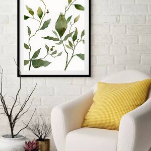 Puede incluir: Una pintura de acuarela enmarcada de hojas y ramas verdes sobre un fondo blanco. La pintura est&aacute; colgada en una pared de ladrillo blanco sobre un sill&oacute;n blanco con un coj&iacute;n amarillo.