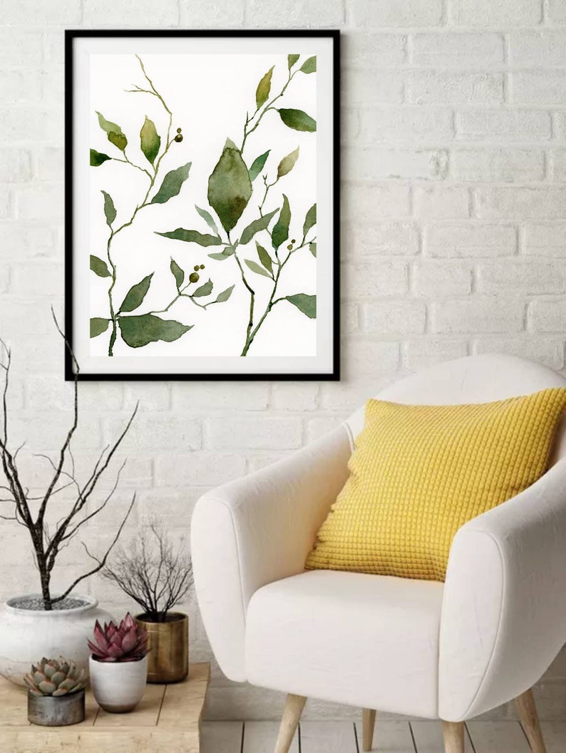 Puede incluir: Una pintura de acuarela enmarcada de hojas y ramas verdes sobre un fondo blanco. La pintura est&aacute; colgada en una pared de ladrillo blanco sobre un sill&oacute;n blanco con un coj&iacute;n amarillo.