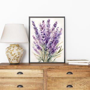 May include: A framed watercolour painting of lavender flowers in shades of purple and green. The artwork is displayed on a wooden chest of drawers next to a decorative lamp with a cream lampshade and a gold floral base.
