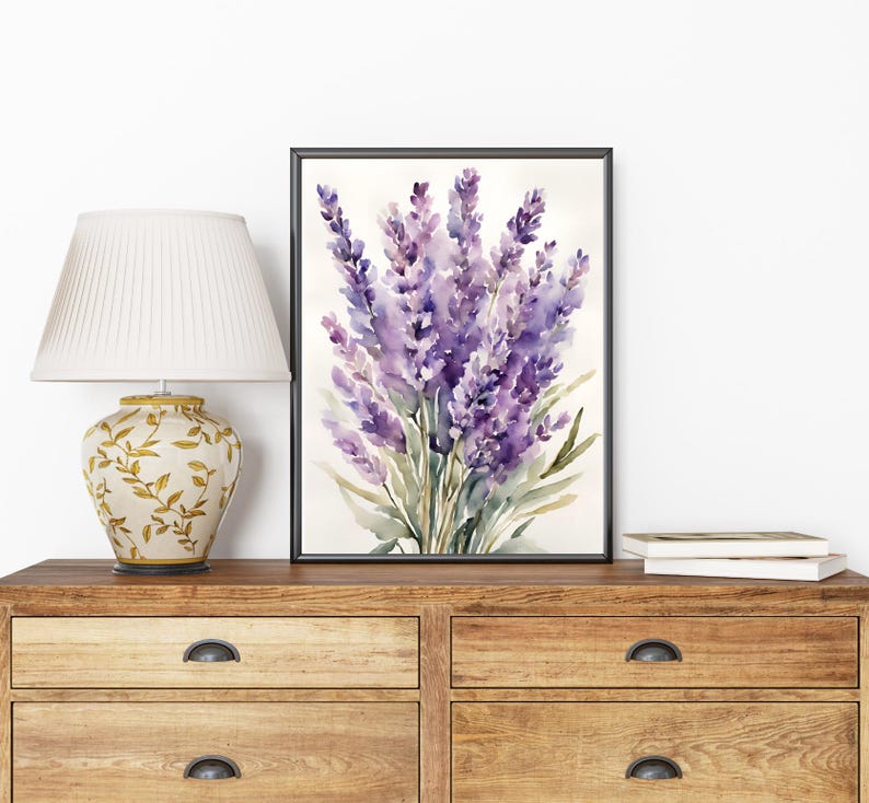 May include: A framed watercolour painting of lavender flowers in shades of purple and green. The artwork is displayed on a wooden chest of drawers next to a decorative lamp with a cream lampshade and a gold floral base.