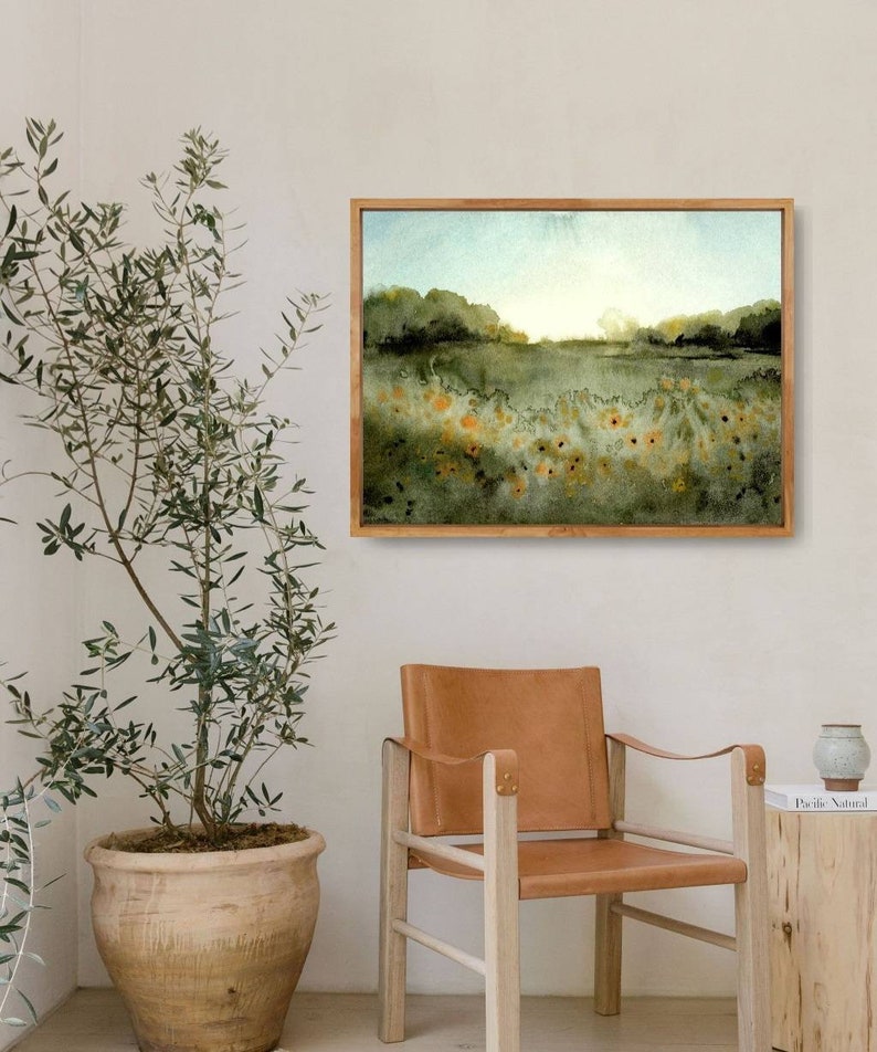 May include: A framed watercolor painting of a field of yellow wildflowers with a green background and a blue sky. The painting is hanging on a white wall above a brown leather chair.