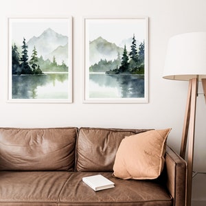 May include: Two framed watercolor paintings of a misty lake scene with mountains and trees reflected in the water. The paintings are in white frames and are hanging on a white wall.