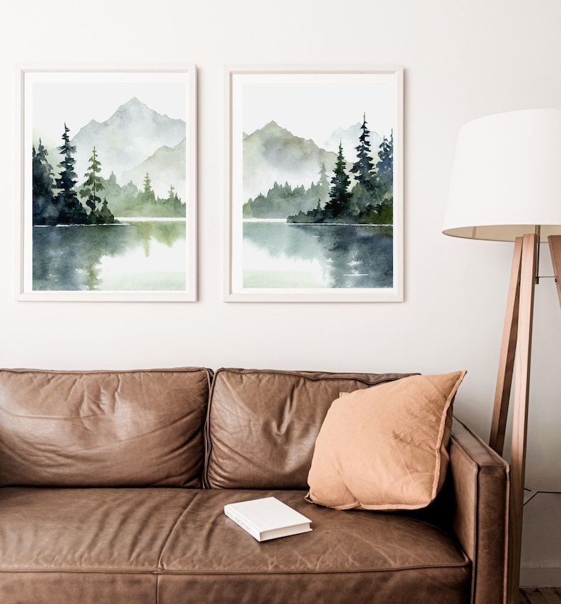 May include: Two framed watercolor paintings of a misty lake scene with mountains and trees reflected in the water. The paintings are in white frames and are hanging on a white wall.