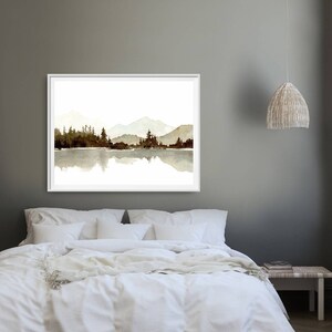May include: A watercolour painting of a mountain range reflected in a lake. The mountains are a muted brown colour and the lake is a light blue. The painting is framed in a white frame and is hanging on a grey wall.
