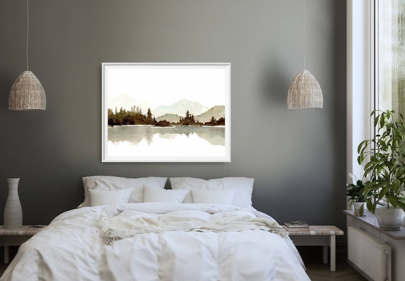 May include: A watercolour painting of a mountain range reflected in a lake. The mountains are a muted brown colour and the lake is a light blue. The painting is framed in a white frame and is hanging on a grey wall.