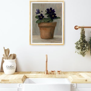 May include: A painting of a potted violet plant with purple flowers in a terracotta pot. The painting is framed in a light wood frame and is hanging on a white wall.