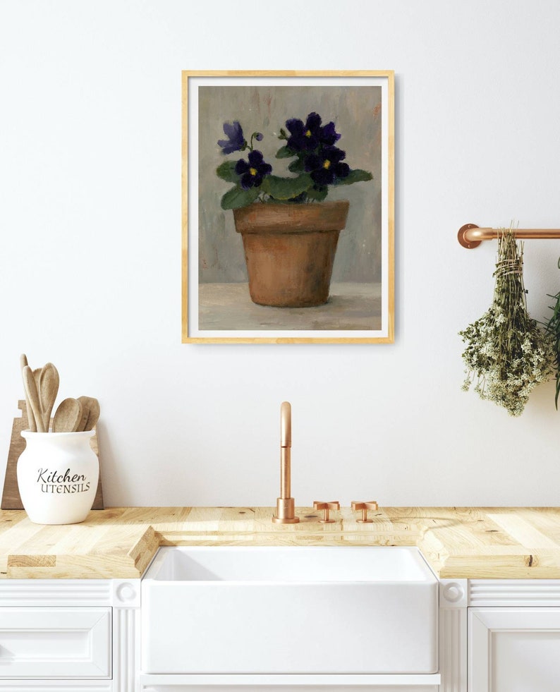 May include: A painting of a potted violet plant with purple flowers in a terracotta pot. The painting is framed in a light wood frame and is hanging on a white wall.