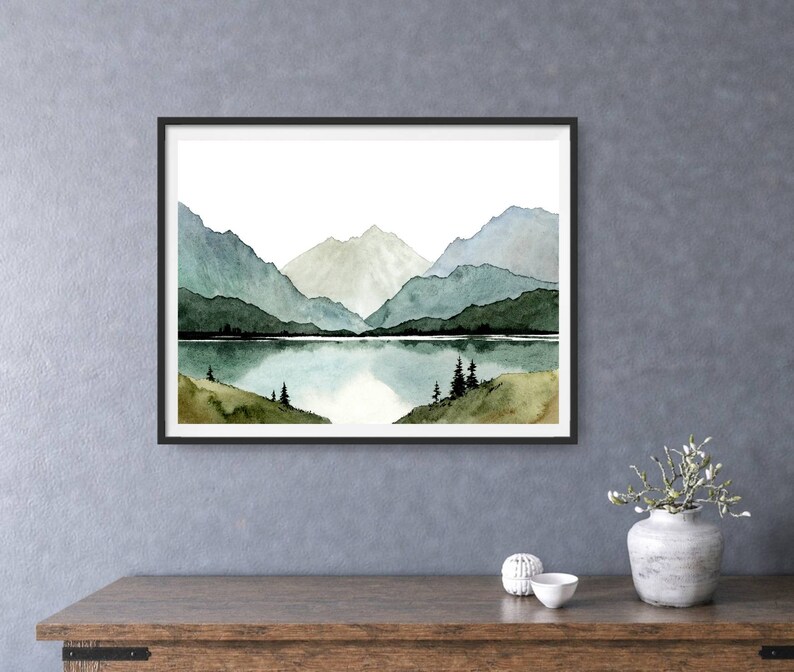 May include: Watercolor painting of a mountain range reflected in a still lake. The mountains are painted in shades of blue and green, with a white peak in the center. The lake is a light blue, and the sky is white.
