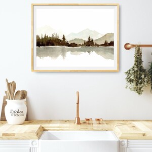 May include: A framed watercolour painting of a mountain range reflected in a lake. The mountains are a light blue and the lake is a light brown. The trees are a dark brown. The painting is in a light wood frame.