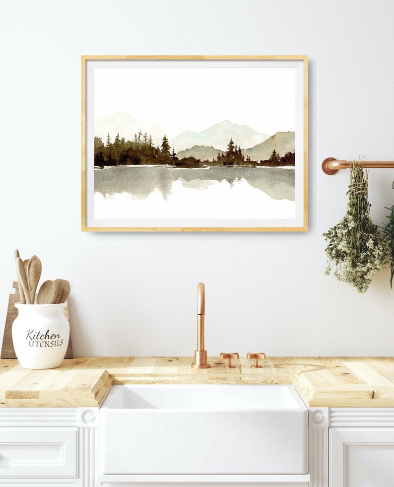 May include: A framed watercolour painting of a mountain range reflected in a lake. The mountains are a light blue and the lake is a light brown. The trees are a dark brown. The painting is in a light wood frame.