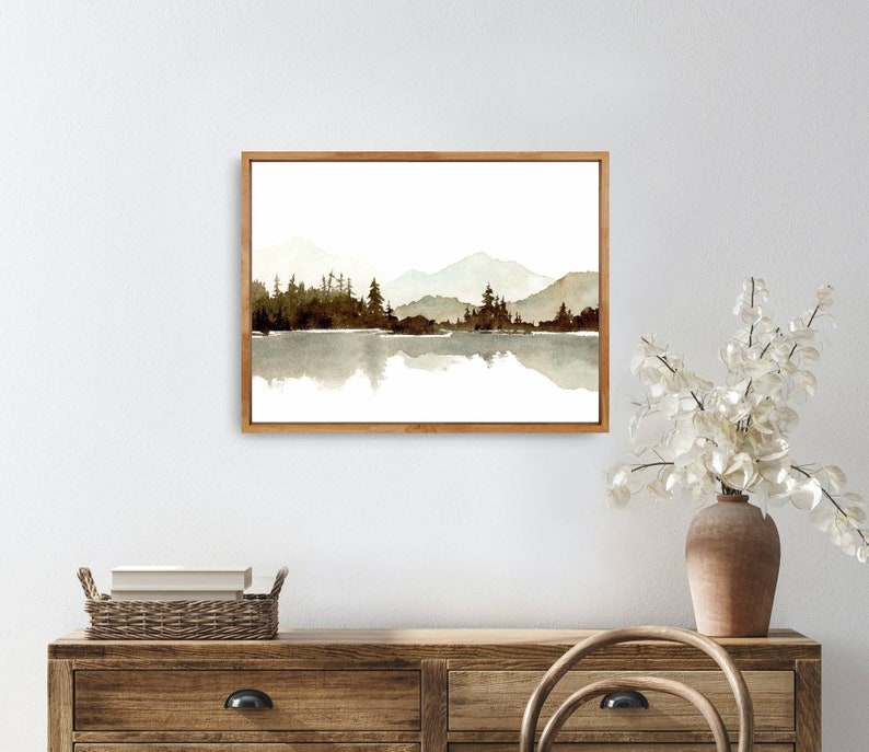 May include: A watercolour painting of a mountain range reflected in a still lake. The mountains are a light brown colour and the lake is a dark brown colour. The painting is framed in a light wood frame.