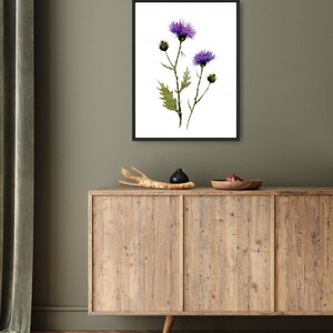 May include: A framed watercolor print featuring purple thistle flowers with green leaves and stems. The artwork is set against a white background within a black frame. The print is displayed on a wooden cabinet with decorative items.
