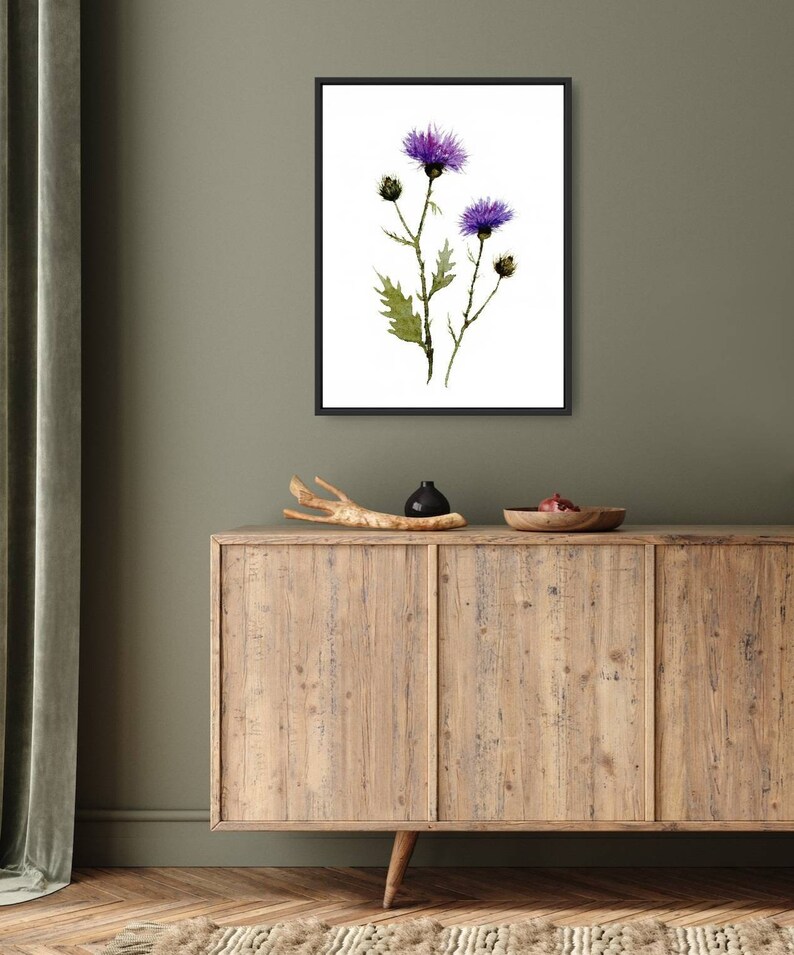 May include: A framed watercolor print featuring purple thistle flowers with green leaves and stems. The artwork is set against a white background within a black frame. The print is displayed on a wooden cabinet with decorative items.