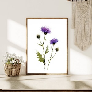 May include: A framed watercolor print featuring purple thistle flowers with green leaves and buds. The artwork is set against a white background, framed in a brown wooden frame. A woven basket of white flowers and a macrame wall hanging are also visible.