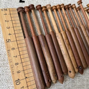 May include: A collection of wooden bobbins in various shades of brown, arranged diagonally. The bobbins have a cylindrical body and a rounded top. A wooden ruler with centimeter markings is placed next to the bobbins. The number "2" is visible in the image.