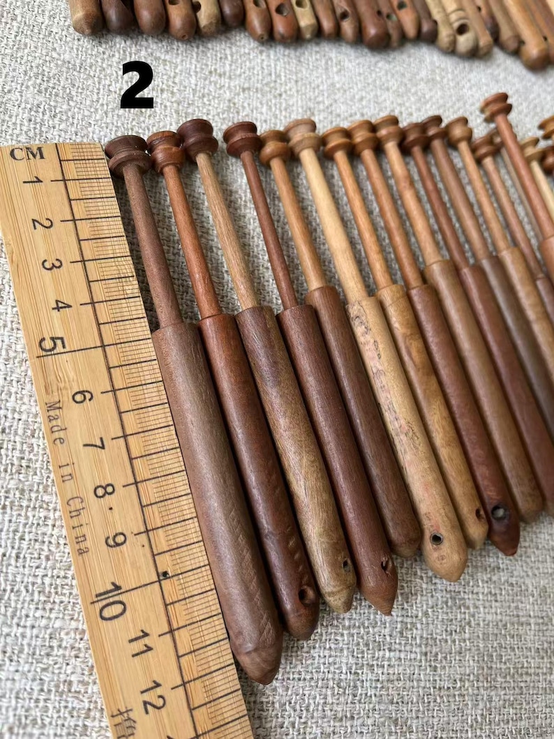 May include: A collection of wooden bobbins in various shades of brown, arranged diagonally. The bobbins have a cylindrical body and a rounded top. A wooden ruler with centimeter markings is placed next to the bobbins. The number "2" is visible in the image.