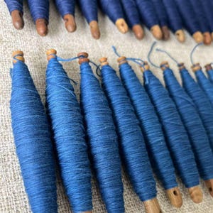 K&ouml;nnte beinhalten: Nahaufnahme einer Gruppe blauer Garnrollen mit Holzenden. Die Rollen sind in einer Reihe angeordnet, wobei die Holzenden zum Betrachter zeigen. Das Garn ist tief indigo-blau.