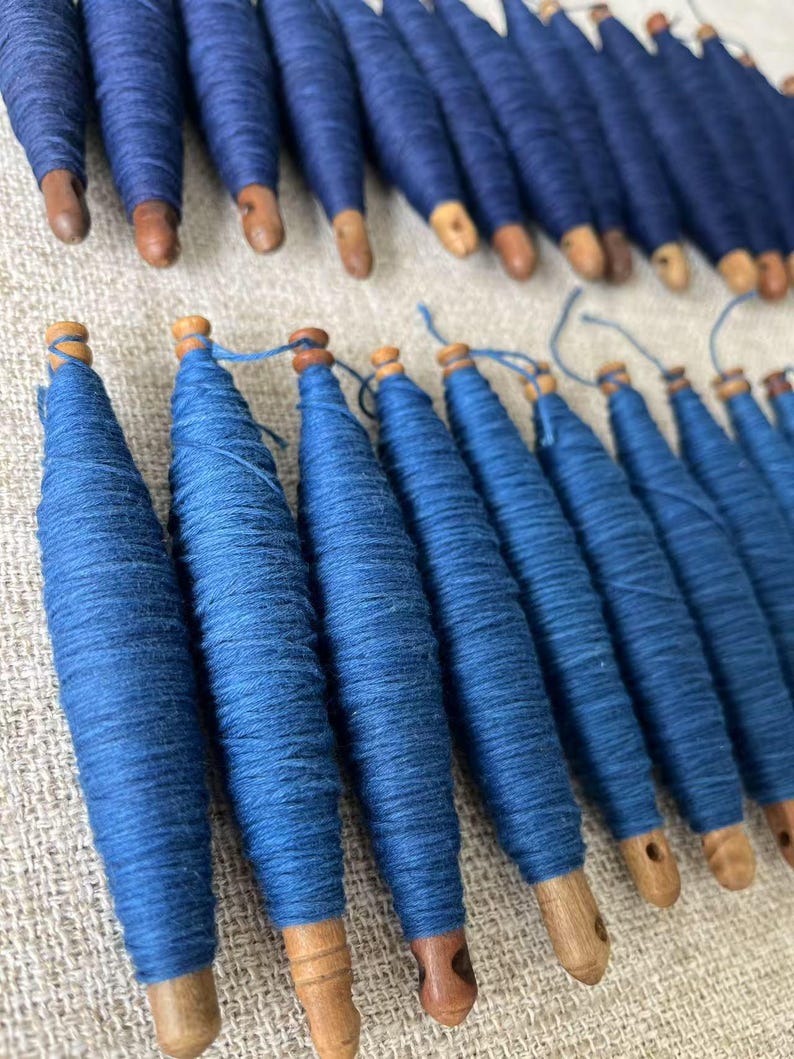 K&ouml;nnte beinhalten: Nahaufnahme einer Gruppe blauer Garnrollen mit Holzenden. Die Rollen sind in einer Reihe angeordnet, wobei die Holzenden zum Betrachter zeigen. Das Garn ist tief indigo-blau.