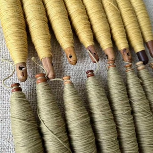 May include: A collection of wooden spools wrapped with yellow and green thread. The spools have a tapered shape and are arranged in rows. The wooden bases are visible, and some have small holes. The thread appears to be tightly wound around the spools.
