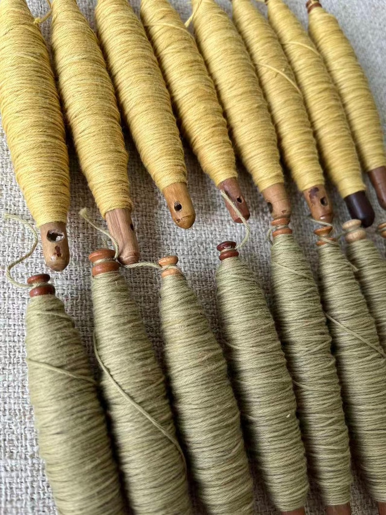 May include: A collection of wooden spools wrapped with yellow and green thread. The spools have a tapered shape and are arranged in rows. The wooden bases are visible, and some have small holes. The thread appears to be tightly wound around the spools.