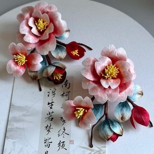 Pode incluir: Dois conjuntos de flores de tecido rosa e vermelho com detalhes azuis e verdes. As flores est&atilde;o dispostas sobre uma superf&iacute;cie branca com um peda&ccedil;o de papel com caligrafia preta.