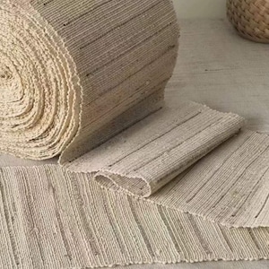 May include: A roll of natural fiber rug material, woven with a striped pattern. The material is unraveled and laid out flat, showing the texture and weave.