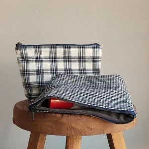 May include: Two fabric zippered pouches with a plaid pattern. The top pouch is white with black and blue plaid. The bottom pouch is black with white gingham. The pouches are sitting on a wooden stool.