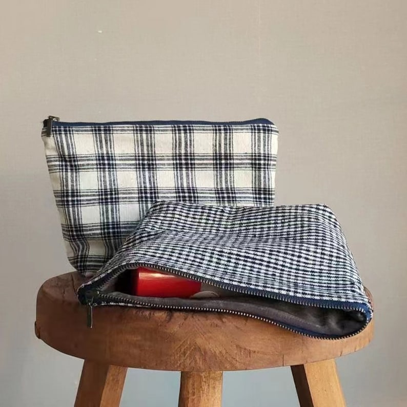 May include: Two fabric zippered pouches with a plaid pattern. The top pouch is white with black and blue plaid. The bottom pouch is black with white gingham. The pouches are sitting on a wooden stool.