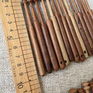 May include: A collection of wooden lace bobbins in various shades of brown, arranged on a textured surface. A wooden ruler with centimeter markings is placed next to the bobbins. The ruler reads "Made in China".