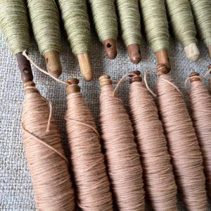 May include: A collection of vintage wooden spools wrapped with thread. The spools are in two colors: light green and a light peach. The wooden bases are visible, and the thread is tightly wound around the spools. The spools are arranged in rows.