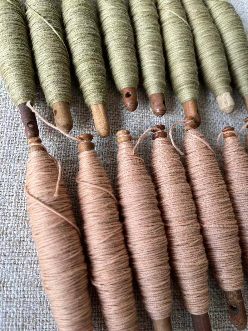 May include: A collection of vintage wooden spools wrapped with thread. The spools are in two colors: light green and a light peach. The wooden bases are visible, and the thread is tightly wound around the spools. The spools are arranged in rows.