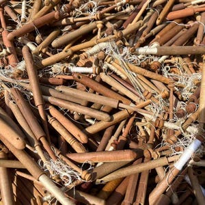 May include: A collection of wooden lace bobbins in various shades of brown, with white thread wound around the spools. The bobbins are of different shapes and sizes, creating a textured, close-up view of the craft tools.
