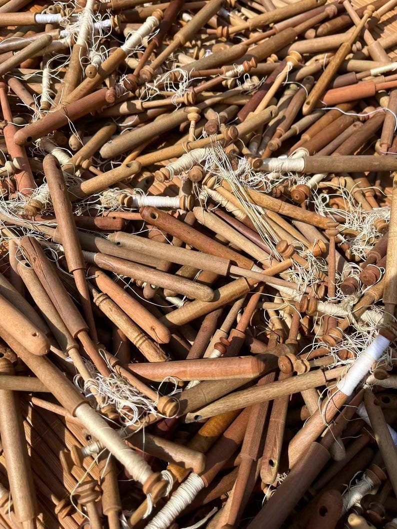 May include: A collection of wooden lace bobbins in various shades of brown, with white thread wound around the spools. The bobbins are of different shapes and sizes, creating a textured, close-up view of the craft tools.